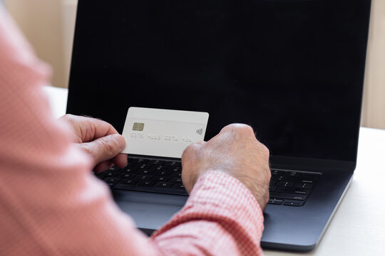 A person in a shirt holds a white bank debit card in front of them against the backdrop of a personal laptop; the shot is taken from over the person's shoulder.