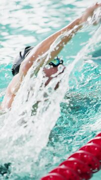 Swimmer in black cap strokes through clear pool water. Red lane divider marks swimmer's path. Water splashes around powerful arm movements. View follows swimmer's back during freestyle stroke