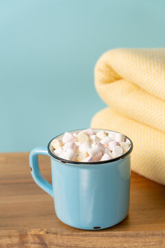 Blue ceramic mug of hot cocoa with marshmallows and a folded yellow blanket on a wooden bench against a soft blue background with copy space