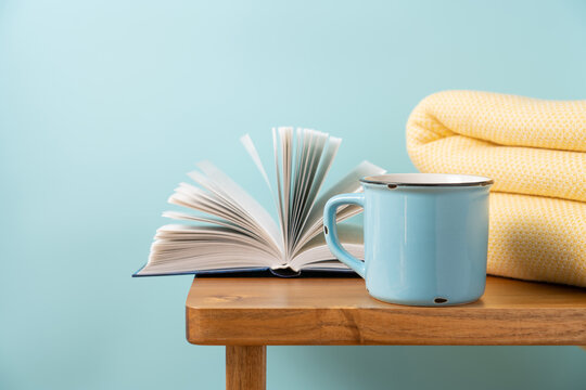 Blue ceramic mug of hot cocoa with a blanket and open book on a wooden bench, cozy home reading and relaxation concept