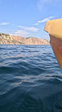 Calm open ocean voyage features bright yellow kayak prow gliding toward distant rugged mountains. First-person perspective tracking shot.