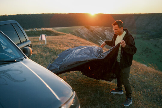 Man assembles a camping tent beside an SUV on a hilltop at sunset, preparing a scenic campsite above a valley and representing road travel, adventure, and outdoor living.