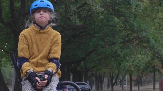A static medium camera shot captures a young girl in a blue helmet and yellow sweater sitting on a park bench taking a peaceful break during roller skating in Kőb&aacute;nya Sportliget Budapest Hungary.