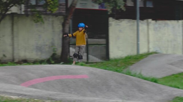 A tracking camera shot follows a kid wearing a blue helmet pumping on an asphalt pumptrack during an outdoor health and sport roller skating activity in Kőb&aacute;nya Sportliget Budapest Hungary.