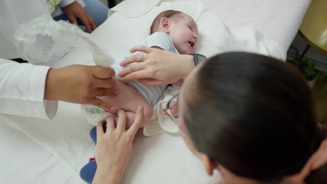High angle view of a medical professional performing a neonatal hip ultrasound screening on a baby while the mother assists and comforts her newborn child in a clinic.