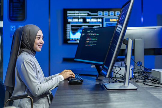 Young Muslim female software developer or programmer smiling while coding on dual computer monitors with lines of code and data dashboards in a modern blue tech office or command center control room.