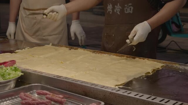 A chef wearing gloves and apron prepares flat savory food on a griddle at an outdoor food stall, with ingredients nearby.