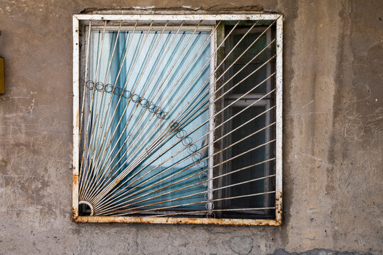 Decorative rusty metal window grille forged in a geometric sunburst pattern and mounted on a weathered concrete wall beside a curtained glass pane, Yerevan, Armenia