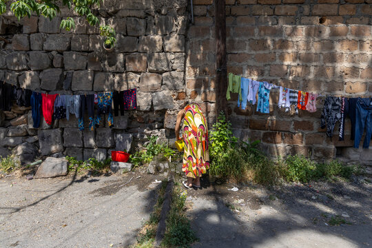 Woman bending over a stone water spring to wash clothes near a wall hung with colorful laundry drying in the shade of a tree in an old courtyard, Yerevan, Armenia