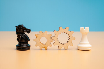 Wooden gears positioned between a black chess knight and a white rook on a beige surface against a blue background. Concept of strategic cooperation, coordinated processes and operational efficiency. © Akmalism Photography