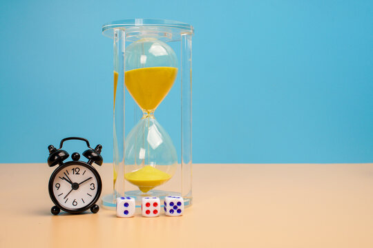 Glass hourglass containing yellow sand beside a black alarm clock and small dice on a beige surface against a blue background. Making strategic decisions before a deadline.