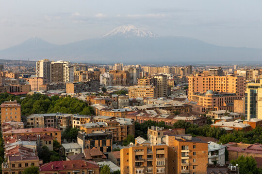 Aerial view of the city skyline with snow capped Mount Ararat rising above rooftops and apartment buildings in the hazy distance at golden hour, Yerevan, Armenia