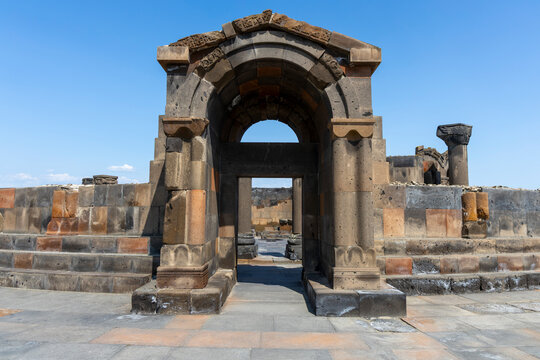 An ancient carved basalt archway with a weathered doorway stands among the sunlit ruins of Zvartnots Cathedral beneath a clear blue summer sky, Etchmiadzin, Armenia
