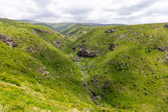 A green rocky canyon carved by a small flowing stream winds through forested hills beneath an overcast cloudy sky in the remote Armenian highlands, Amberd, Armenia
