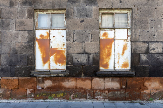 Boarded up windows covered with rusted metal sheets mark the weathered facade of an abandoned black stone building showing signs of urban decay, Gyumri, Armenia