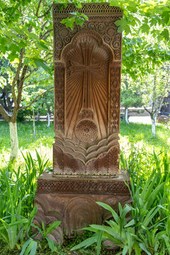 A carved khachkar cross stone stands amid green grass and leafy trees in a quiet sunlit garden filled with soft spring greenery and gentle shade, Gyumri, Armenia