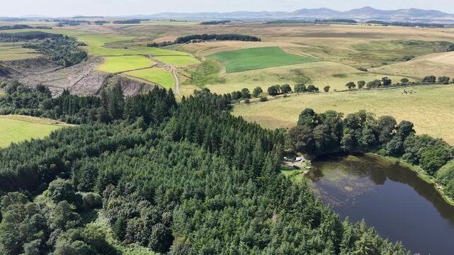 A sweeping aerial drone shot over the vibrant green fields surrounding Edgelaw Reservoir in Midlothian, Scotland, on a bright, sunny summer's day. 
