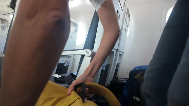 Woman passenger stowing yellow backpack in airplane overhead compartment. Female traveler placing hand luggage in bin before departure. Safe journey and travel essentials