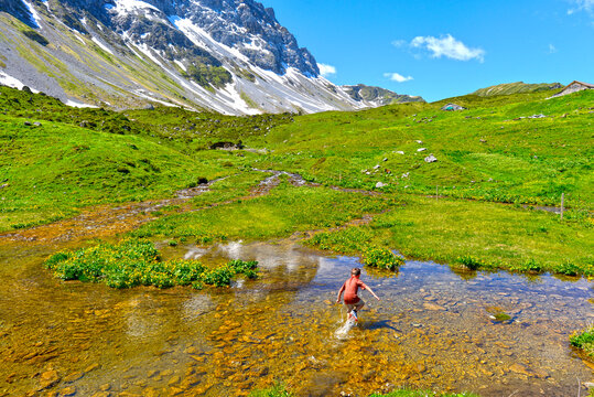 Untersch&auml;chen, Oberalp, Kanton Uri, Schweiz: Kind rennt und springt durch einen flachen Gebirgsbach auf einer gr&uuml;nen Almwiese vor einer schneebedeckten Bergkulisse