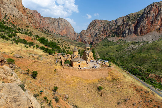An elevated aerial view shows the medieval Surb Astvatsatsin monastery complex with stone walls and a road surrounded by dramatic red rock cliffs, Noravank, Armenia