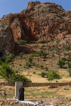 A priest in a black robe prays with his back turned before a newly carved khachkar monument standing near a stone wall close to the Noravank monastery, Noravank, Armenia
