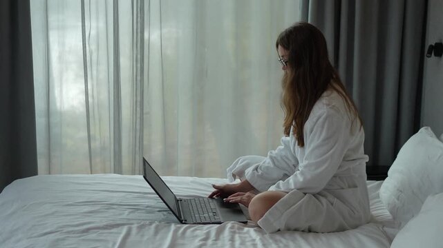 Side view of a female freelancer sitting on a bed and working on a laptop in a modern minimalist bedroom.