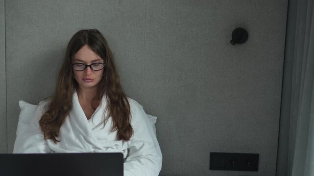 Focused young woman in a white bathrobe working on a laptop while sitting in a hotel bed with a static medium shot.