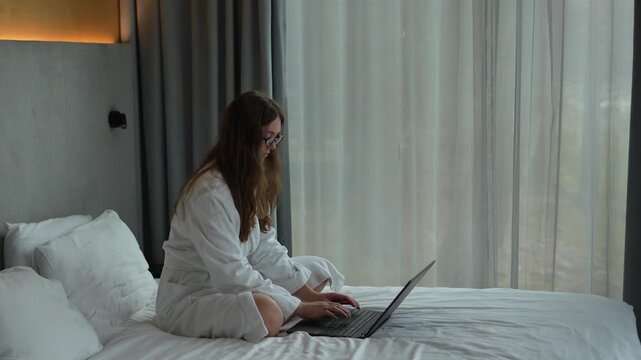 Young woman in a white bathrobe sitting cross-legged on a hotel bed and using a laptop on a sunny morning.