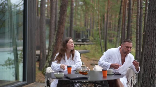 Funny young man in a white bathrobe stuffing a whole cupcake into his mouth during a breakfast at a forest resort terrace.