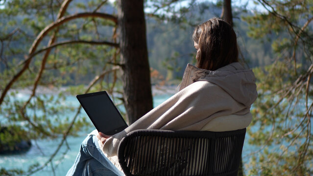 Back three-quarter view of a female tourist wrapped in a warm blanket using a digital tablet while relaxing on an outdoor terrace near a turquoise river.