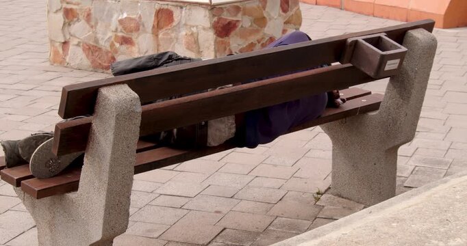 Poor homeless man or refugee sleeping on the wooden bench on the urban street in the city.