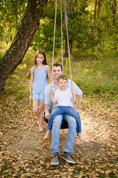 Happy father with son and daughter playing on tire swing in autumn park. Cute boy and older sister having fun with dad on rope swing outdoors. Active fatherhood, carefree childhood concept, parenting