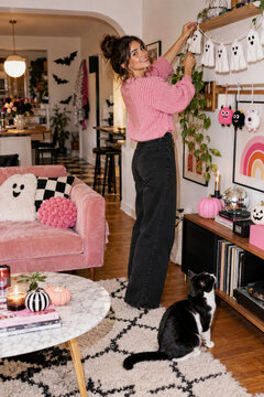 Woman hanging playful ghost garland in a pink Halloween apartment with black cat