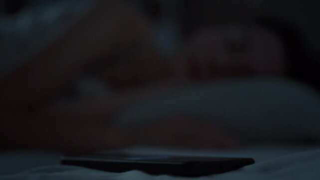 An incoming call lights up a smartphone in a dark bedroom. A sleeping young woman under a blanket reaches for the phone instinctively. Concept of sleep disruption and always-connected digital life