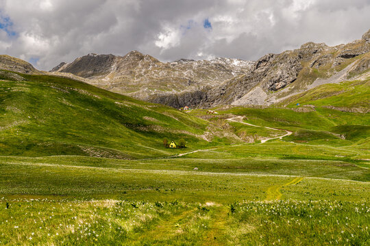 Panoramic view of a lush alpine valley with winding road, remote cabins and rugged peaks with lingering snow. Dramatic clouds and green meadows create a scenic destination for hiking and mountain