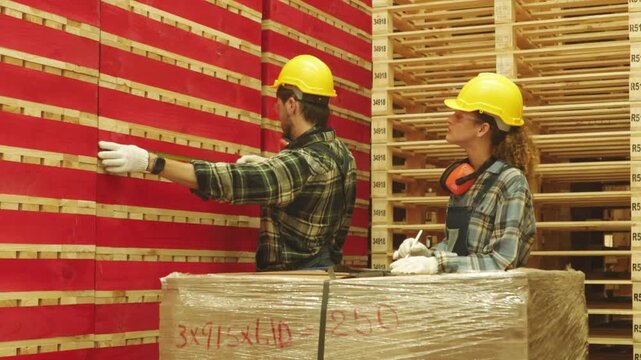 Male and female carpenter team wearing hard hats and safety glasses measure red painted pallet lumber tape measure the organized stacking handling these materials industrial facility classification.