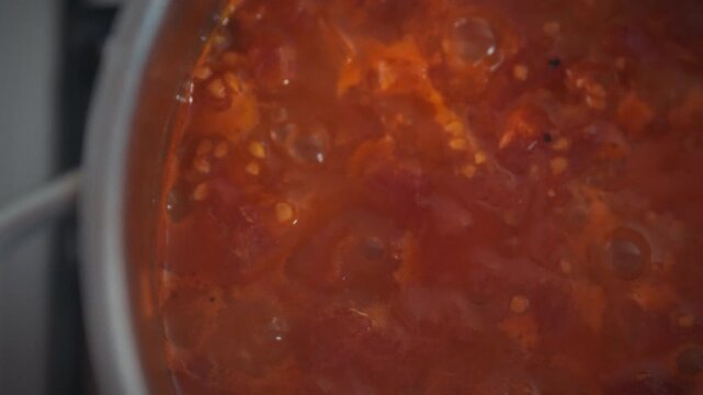 Close-up top view of tomato sauce bubbling in a metal pot on the stove. Chunks of tomato pulp visible in the boiling sauce. Theme of homemade cooking and natural food preparation
