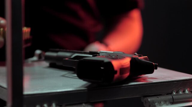 Close-up view of hand gripping handgun on metal table with fingers tight on grip under red moody studio lighting, dark shadows and intense focus