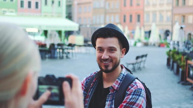 A blonde woman photographs a smiling man wearing a fedora hat in a busy European market square with outdoor seating and multi-story buildings during daytime.