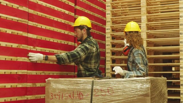 Male and female carpenter team wearing hard hats and safety glasses measure red painted pallet lumber tape measure the organized stacking handling these materials industrial facility classification.