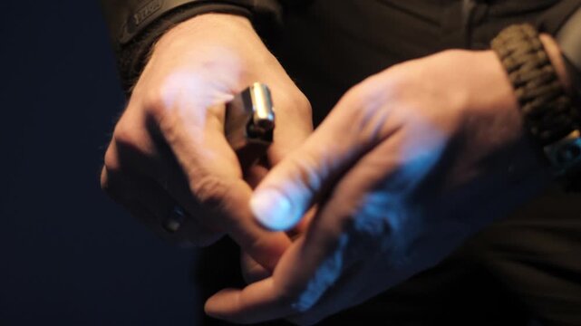 Close-up hands manipulating small pistol while sliding gold ring onto finger in dim studio with warm skin tones and blue glow