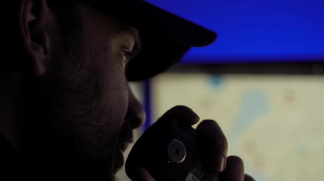 Close-up of cap-wearing man speaking into handheld radio, side profile in dark control room, blue backlight illuminating face and radio equipment with blurred monitors behind