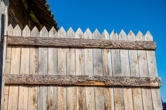 Pointed wooden fence pickets with rough bark crossbeams form a rustic gate beneath a clear blue sky.