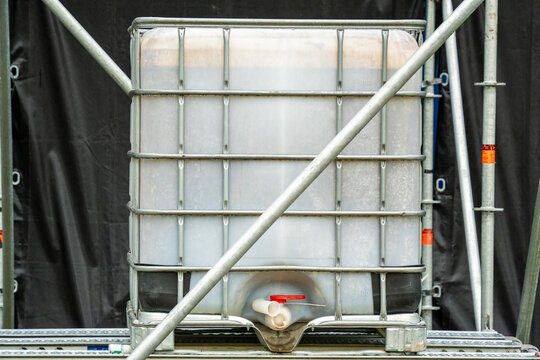 A plastic intermediate bulk container IBC tote tank enclosed in a protective steel cage frame sits on a stage structure in front of black tarpaulin backdrop.