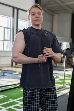 Young athletic man standing inside bright gym near turf training area, wearing sleeveless black shirt and checkered shorts, hands clasped at waist with visible forearm muscles and relaxed confident