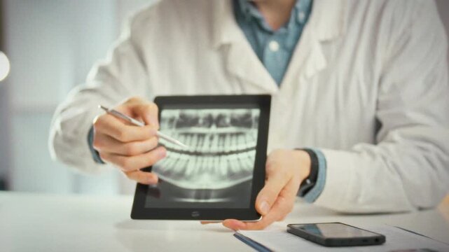 Examining patient's X-ray, doctor in crisp white coat sits at a table in a bright medical office. Using a tablet, he highlights problem areas and discusses tailored treatment options with the patient