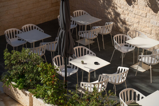 Spacious courtyard with multiple outdoor dining tables and white chairs, surrounded by stone walls, flowering planters and closed umbrellas