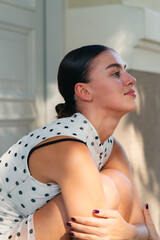 Young brunette woman in a white polka dot dress hugging her knees and looking up in profile, thoughtful outdoor portrait. © Vika