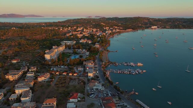 Drone view of the beachfront AKS Hinitsa Bay Hotel in Porto Heli, Greece, with waterfront villas, boats scattered across the calm bay, and warm sunset light glowing overhead.