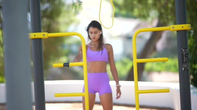Young woman prepares between outdoor calisthenics parallel bars, wearing lilac activewear, pausing to look down at her smartwatch and hand before starting workout